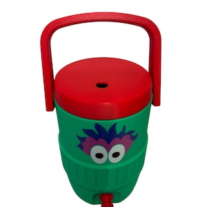 May include: A green plastic beverage cooler featuring a red lid and a red carrying handle. The front of the cooler displays a graphic of a purple and pink creature with large white eyes. A small red spigot is located at the bottom of the cooler.