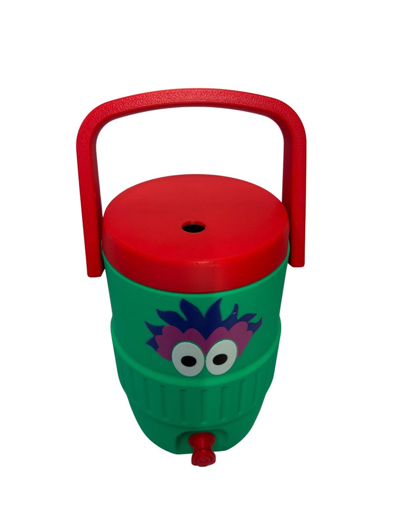 May include: A green plastic beverage cooler featuring a red lid and a red carrying handle. The front of the cooler displays a graphic of a purple and pink creature with large white eyes. A small red spigot is located at the bottom of the cooler.