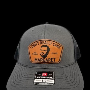 May include: A gray and black trucker hat with a brown leather patch that reads "I don't really care, Vice President, JD Vance, Margaret". The hat is adjustable and has a Richardson 112 logo.
