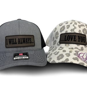 May include: Two trucker caps. The left cap is heather grey with black mesh and a rectangular patch reading I WILL ALWAYS. The right cap features a white and grey leopard print with a white mesh back and a rectangular patch reading ...LOVE YOU.