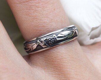 May include: Silver ring with an engraved floral design. The ring features detailed carvings of leaves and flowers around the band. The ring is worn on a finger, with a white flower in the background.