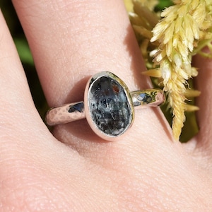 May include: A silver ring with a dark blue-green, oval-shaped stone. The ring's band has a hammered texture. The ring is displayed on a hand, with a blurred background of yellow flowers and greenery.