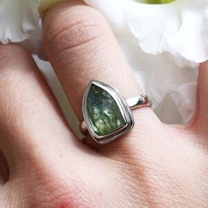 May include: A silver ring with a green gemstone set in a teardrop shape. The ring is on a person's finger.