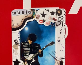 May include: A decorative white framed image featuring a musician playing a white guitar. The frame is adorned with stickers of stars, lips, cherries, and the word "music". The word "heals" is also visible. The background is red.