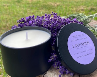 May include: A black candle jar with a white soy wax candle and a lavender scent. The jar has a black lid with the text "Fairy Greens,  Elliott City, MD, Natural Soy Wax, Lavender, Hand Poured Candle, FairyGreens.com". The candle is surrounded by purple lavender flowers.