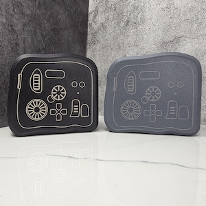 May include: Two gaming console-shaped cases, one black and one gray, with white controller button designs. The cases have a rounded rectangular shape and a textured surface. The controller designs include a directional pad, buttons, and a wheel.