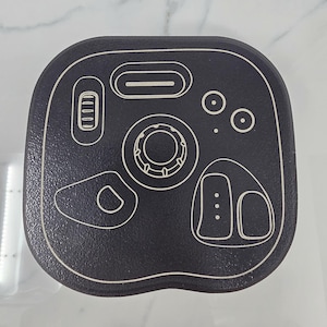 May include: A black, rounded-corner square object with white line art. The design includes a central circular element, a long oval, a battery indicator, and other geometric shapes. The surface has a textured appearance.