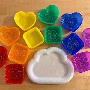 May include: A set of 10 small heart and square shaped resin dishes with glitter embedded in the resin. The dishes are in various colors including red, orange, yellow, green, blue, and purple. There is also a white cloud shaped dish.