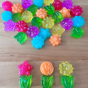 May include: A collection of colorful, glittery flower-shaped resin ornaments. The ornaments are in various colors, including pink, yellow, green, blue, and purple. There are also three larger ornaments in the shape of a flower with a green stem.