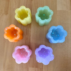 May include: Six flower-shaped candle holders in various pastel colors, including yellow, green, orange, pink, and purple. Each holder has a small, round opening in the center and is decorated with glitter.