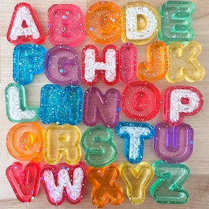May include: A set of 26 colorful, translucent alphabet letter molds filled with white rice. Each letter is a different color and has a glittery finish.