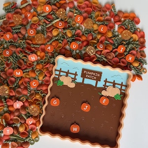 May include: A square tray with a wavy edge featuring a pumpkin patch illustration and the text PUMPKIN PATCH. The tray contains embossed letters and orange pumpkin-shaped markers. It is surrounded by loose orange and green pasta shapes.