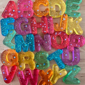 May include: A set of 26 colorful, translucent alphabet letter tiles with glitter embedded in the plastic. The letters are arranged in alphabetical order.