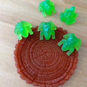 May include: Four green frog figurines with glitter accents sit on a brown tree stump-shaped dish. The frogs are arranged around the edge of the dish, with their heads facing inward.