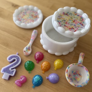 May include: A white cake-shaped box with a lid decorated with colorful sprinkles. The box is open and the lid is sitting next to it. There are also several small, colorful balloons, a purple number two candle, and two spoons decorated with sprinkles.