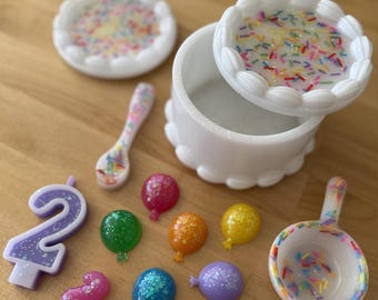 May include: A white cake-shaped box with a lid decorated with colorful sprinkles. The box is open and the lid is sitting next to it. There are also several small, colorful balloons, a purple number two candle, and two spoons decorated with sprinkles.