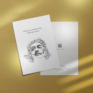 May include: Two white greeting cards with a gold background. The front card features a black and white illustration of a man's face with a mustache and the text "Cheers to another year, of being surreal."