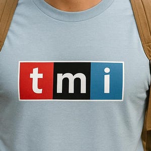 May include: A light blue t-shirt with the letters "tmi" in a rectangular design. The "t" is red, the "m" is black, and the "i" is blue. The person is wearing a green beanie, glasses, and a backpack.