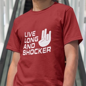 May include: A maroon t-shirt with the text "LIVE LONG AND SHOCKER" in white, along with a hand gesture. The shirt is worn with navy blue pants and a white watch. The person is wearing sunglasses.