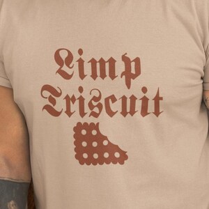 May include: Beige t-shirt with the text "Pimp Triscuit" in a burgundy gothic font and a matching illustration of a cracker. The shirt is a short-sleeved crew neck.