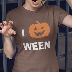 Puede incluir: Camiseta marrón con el texto "I WEEN" y un diseño de calabaza naranja. La camiseta es de manga corta y cuello redondo. La persona lleva vaqueros oscuros.