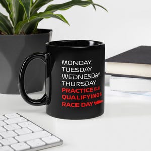 Puede incluir: Taza de café de cerámica negra con texto blanco que dice "Lunes, Martes, Miércoles, Jueves, Práctica, Clasificación, Día de carrera" en rojo. Un pequeño gráfico de coche de carreras rojo está al lado del texto "Día de carrera".