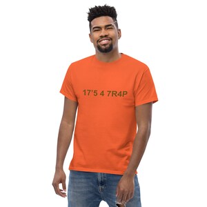 Peut inclure: Un homme portant un t-shirt orange avec le texte "17'5 4 7R4P" imprimé en marron sur le devant.