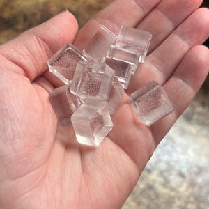 May include: A handful of clear, square glass tiles with a subtle glitter effect. The tiles are held in an open palm, showcasing their translucent quality and geometric shape. The tiles appear to be for crafting or decorative purposes.