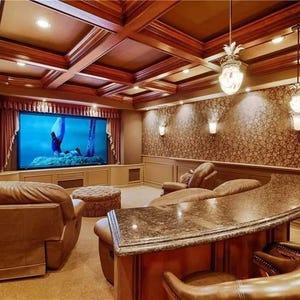 May include: A home theater room with a large screen displaying a movie. The room features brown leather seating, a curved bar with stools, and decorative lighting. The ceiling is made of wood.