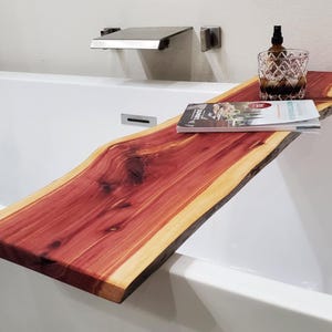 Puede incluir: Una bandeja de baño de madera natural larga con un tono marrón rojizo. La bandeja se coloca sobre una bañera blanca y tiene un libro y una botella de vidrio en la parte superior.