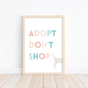 Peut inclure: Une impression encadrée avec le texte "ADOPTER NE PAS ACHETER" en rose pastel, bleu et vert. Une petite silhouette grise de la queue et des pattes arrière d'un chien est visible sur le côté droit de l'impression.