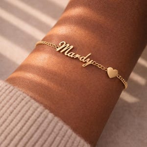 May include: A gold-colored bracelet with the name "Mardy" in cursive script. A small heart charm is attached to the bracelet. The bracelet is worn on a wrist, with a neutral-colored sweater in the background.
