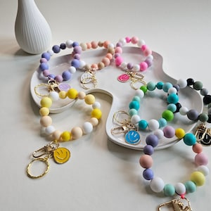 Könnte beinhalten: Ein Set aus bunten Perlenarmbändern mit Smiley-Anhängern. Jedes Armband besteht aus Perlen in verschiedenen Farben und hat einen goldenen Schlüsselring.