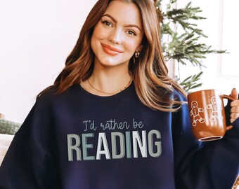 May include: Navy blue sweatshirt with the text "I'd rather be READING" in a gradient font. A brown mug with gingerbread men is held in the hand. A plush deer pillow and decorative houses are in the background.