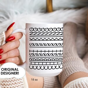 May include: A white 15 oz coffee mug with a series of black geometric patterns. The mug is held by a person with red nail polish. The words "Original Designer!" are in a white cloud-shaped text box.