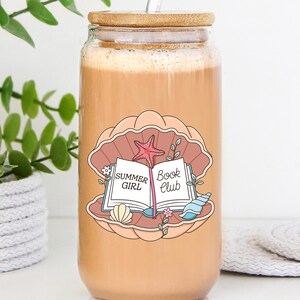 May include: A glass tumbler with a wooden lid and a clear straw, filled with a light brown beverage. The tumbler features a seashell design with a book and the words "Summer Girl Book Club".