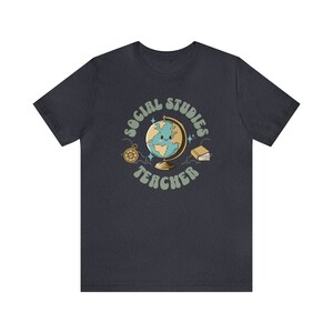 May include: Dark blue t-shirt with a retro-style graphic of a globe with a smiling face, surrounded by stars, a compass, and a book. The text "Social Studies Teacher" is written in a retro font around the globe.