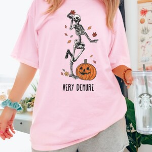 May include: A pink t-shirt with a skeleton graphic holding a leaf and standing on a jack-o-lantern. The text "VERY DEMURE" is printed below the graphic.