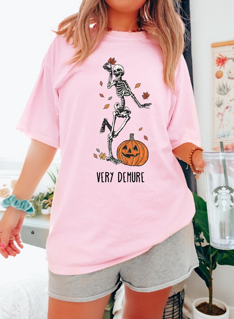 May include: A pink t-shirt with a skeleton graphic holding a leaf and standing on a jack-o-lantern. The text "VERY DEMURE" is printed below the graphic.