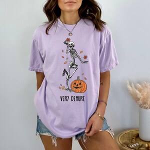 May include: A lavender t-shirt with a graphic of a skeleton dancing on a pumpkin with the text "Very Demure".