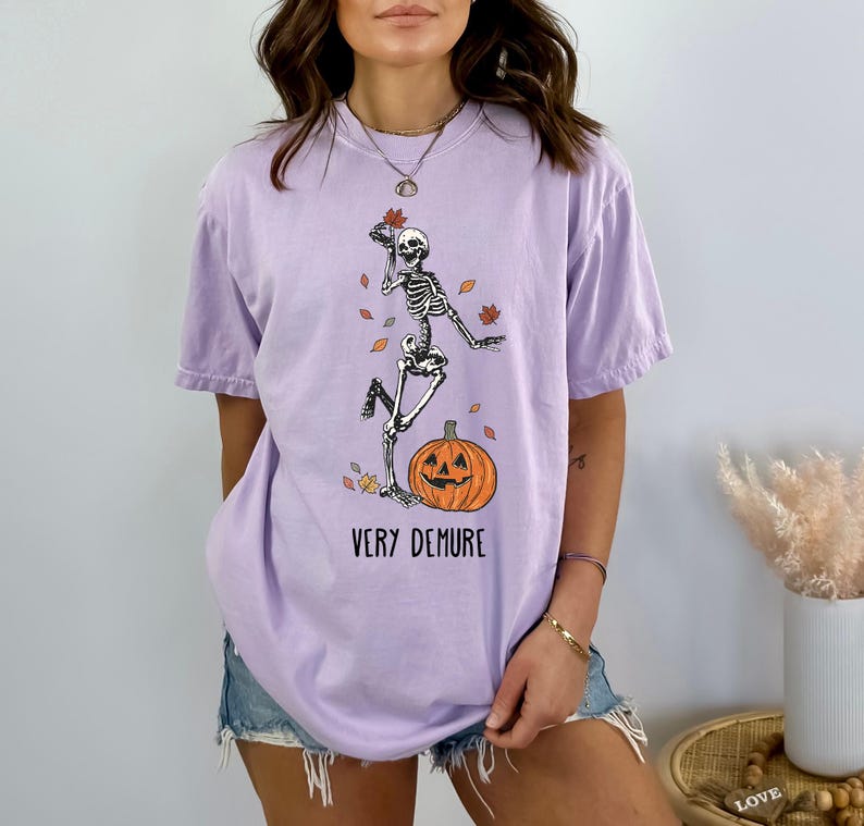 May include: A lavender t-shirt with a graphic of a skeleton dancing on a pumpkin with the text "Very Demure".