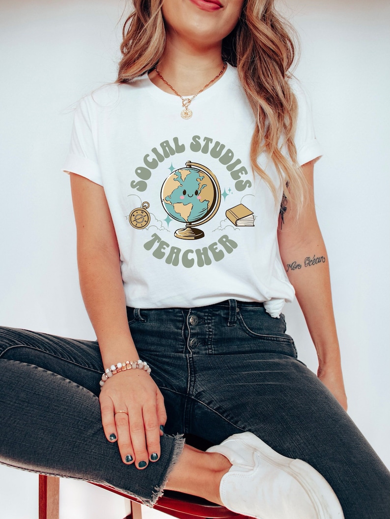 May include: White t-shirt with a graphic of a globe with a smiling face, surrounded by a circle that says "Social Studies Teacher" in green letters.