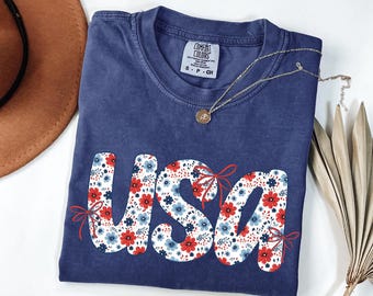 May include: A navy blue t-shirt with the word "USA" in a floral design of red and blue flowers. The shirt has a Comfort Colors tag and is paired with a gold necklace.