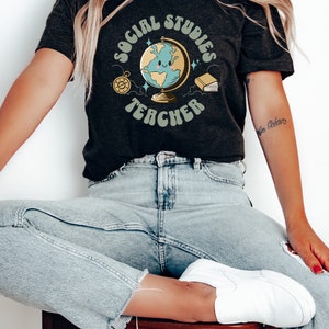 May include: A dark grey t-shirt with the text "Social Studies Teacher" in a retro font. The design features a cartoon globe with a smiling face, a compass, and a book.
