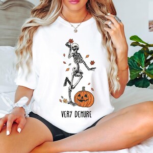 May include: White t-shirt with a skeleton dancing next to a jack-o'-lantern. The text "Very Demure" is printed below the image.