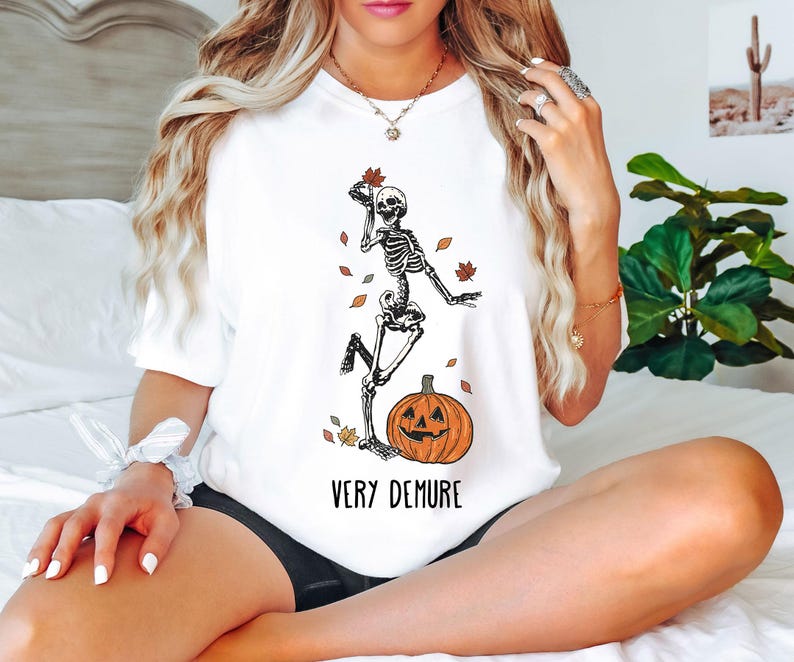 May include: White t-shirt with a skeleton dancing next to a jack-o'-lantern. The text "Very Demure" is printed below the image.