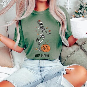 May include: A green t-shirt with a skeleton dancing on a pumpkin. The text "Very Demure" is printed below the image.