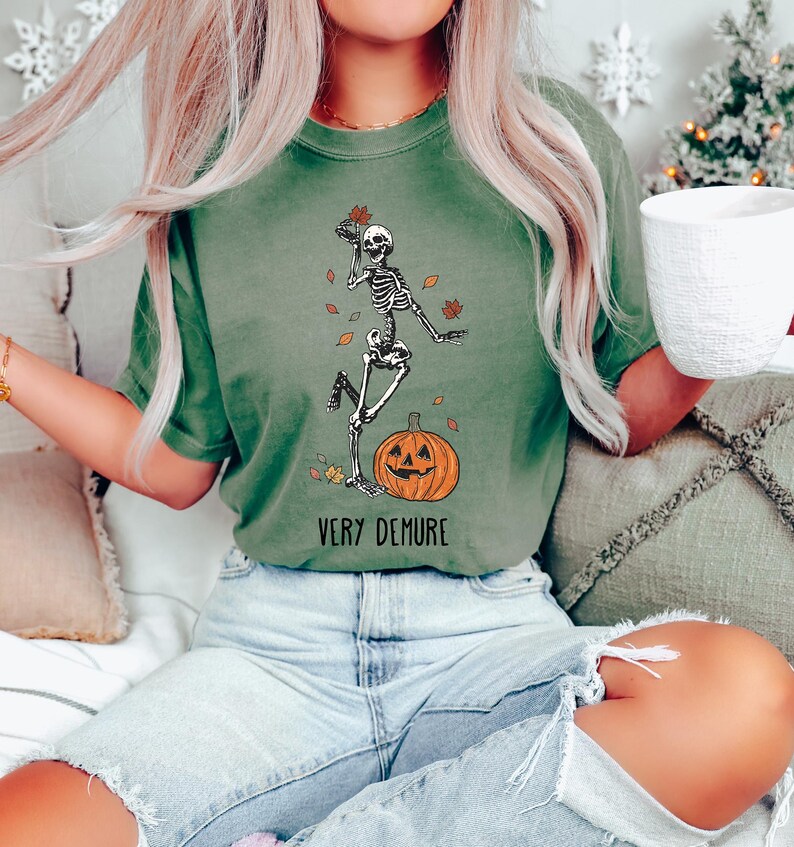 May include: A green t-shirt with a skeleton dancing on a pumpkin. The text "Very Demure" is printed below the image.