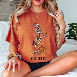 May include: A burnt orange t-shirt with a skeleton dancing on a pumpkin. The text "Very Demure" is printed on the bottom of the shirt.