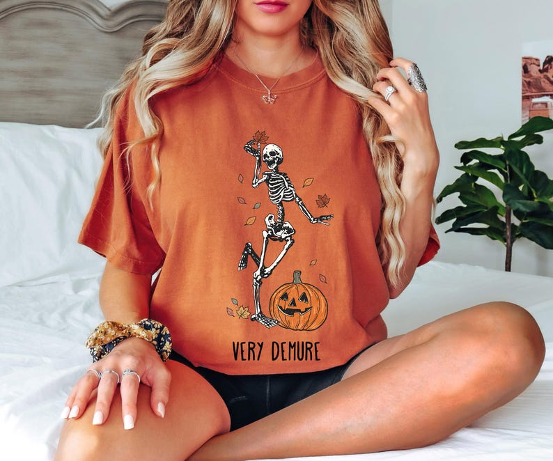 May include: A burnt orange t-shirt with a skeleton dancing on a pumpkin. The text "Very Demure" is printed on the bottom of the shirt.